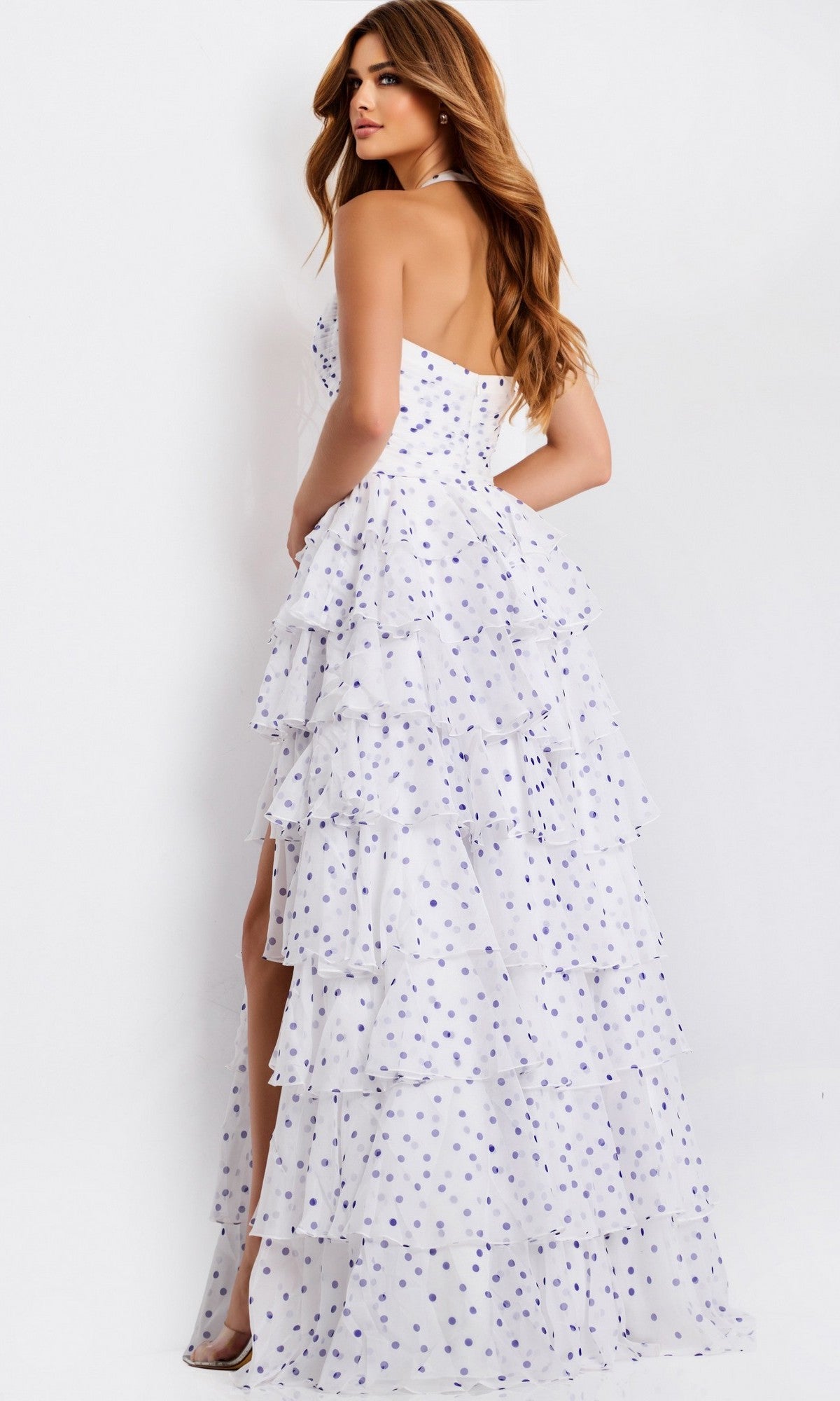 A woman in the Jovani 49776 long prom dress, featuring white polka dots, tiered ruffles, and a sweetheart halter neckline, stands sideways and looks over her shoulder against a plain background.